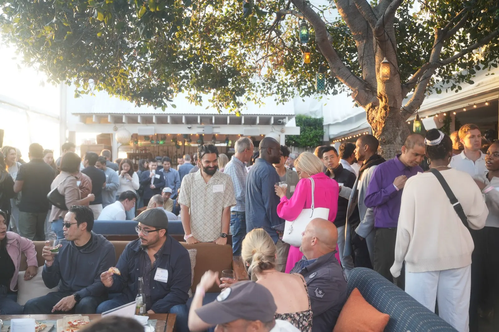 The Bungalow patio at dusk, packed — founders and operators talking in clusters under the fig tree, drinks in hand, the bar and string lights glowing behind them.