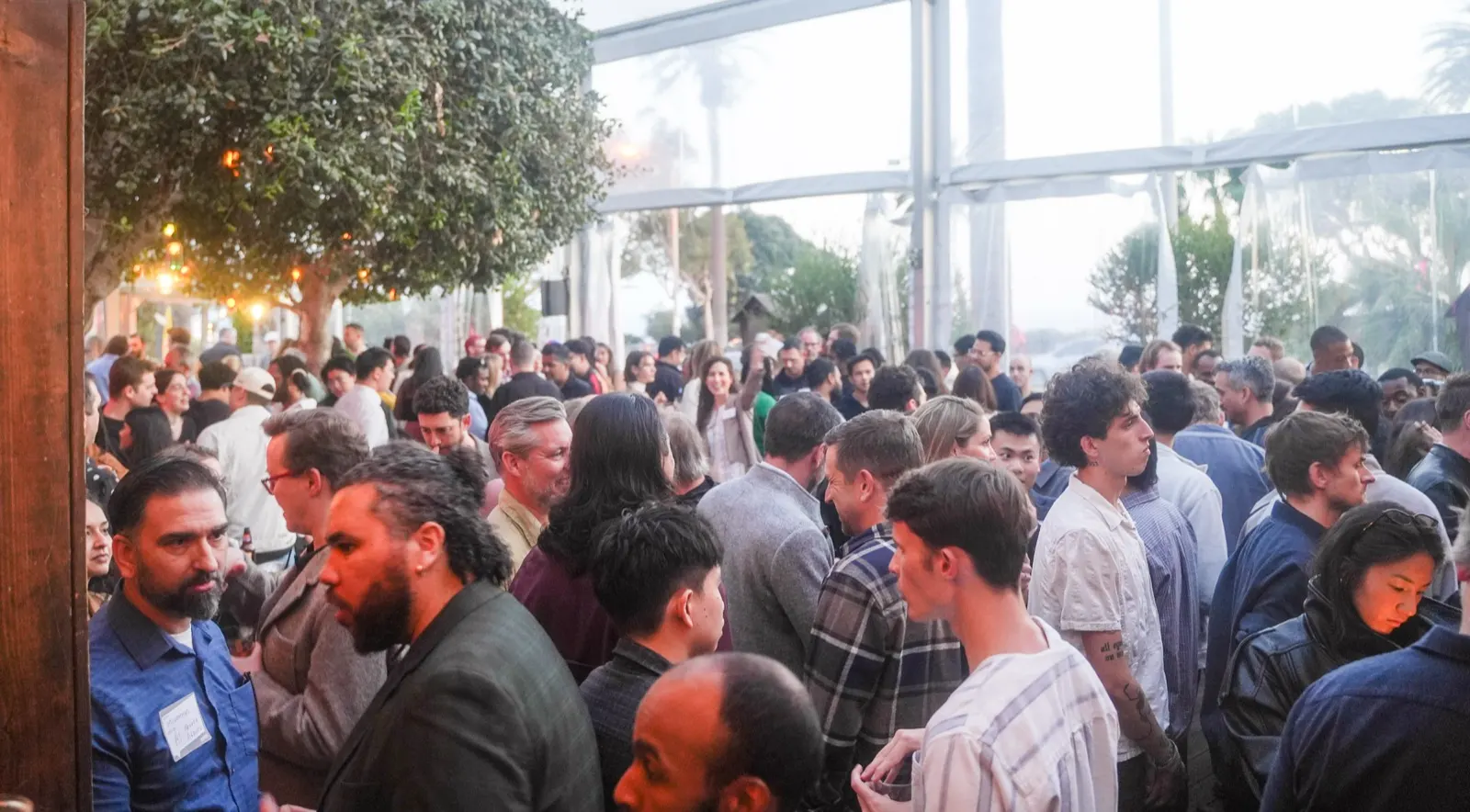A wall-to-wall crowd inside the tented room at The Bungalow, name tags on, the patio and palms visible through the clear walls.