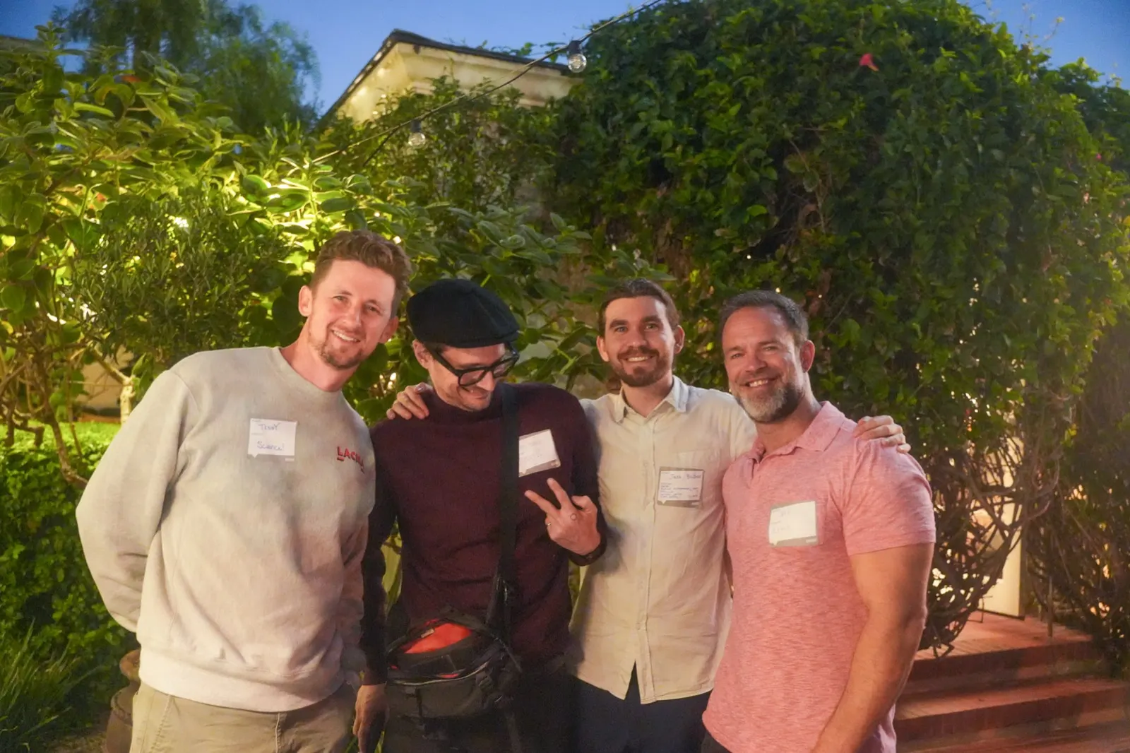 Four founders arm in arm for a photo in front of the ivy wall, lit warm against the evening.