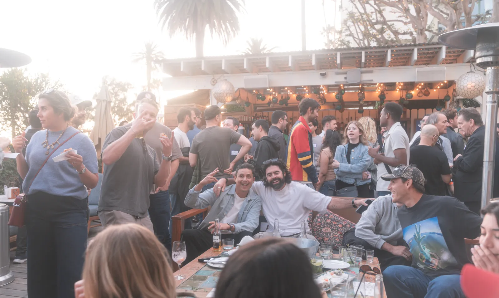 A packed patio at The Bungalow — the crowd filling the tables and bar under string lights and woven pendant lamps, two guests grinning at the camera.