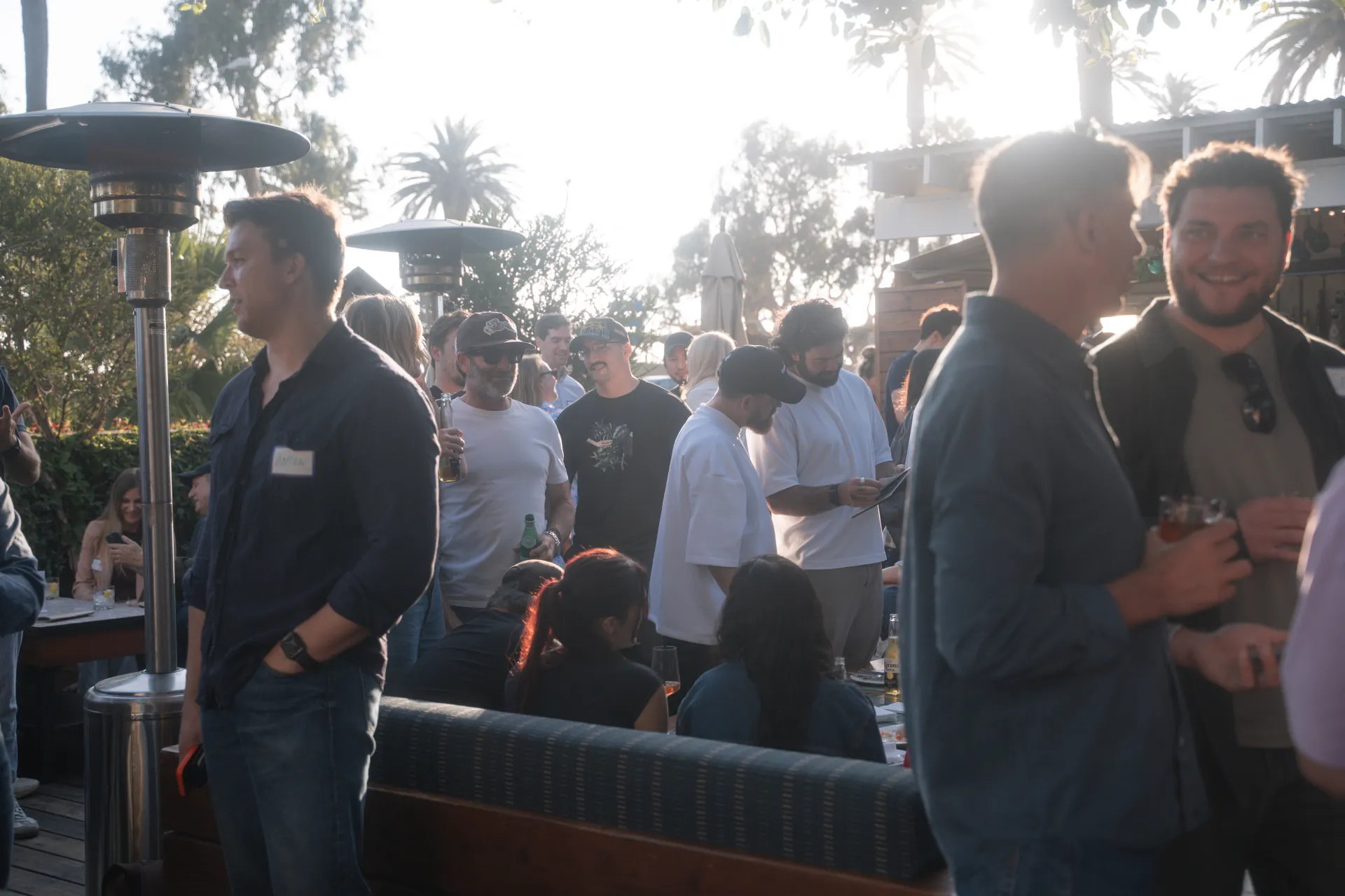Founders and operators with drinks in hand on The Bungalow's open patio at golden hour, palms backlit by the low sun behind them.