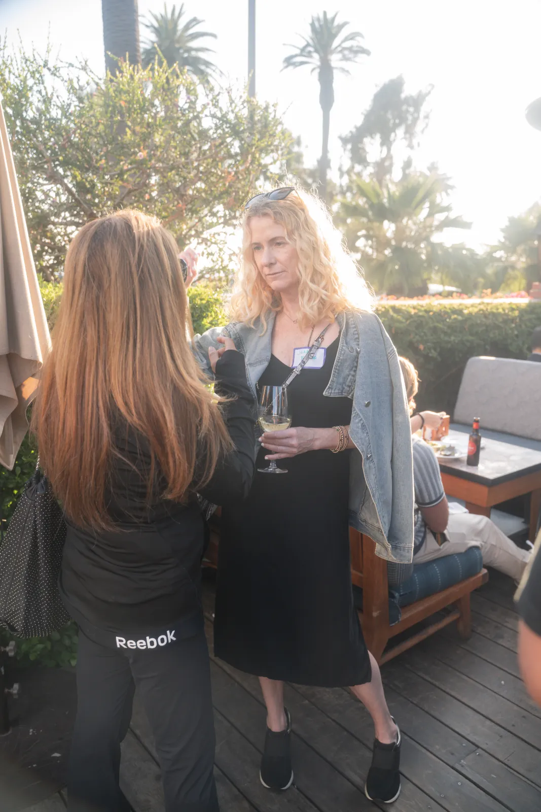 Two attendees talking over a glass of wine on the wood deck, palms and hedges glowing in the late-afternoon light.