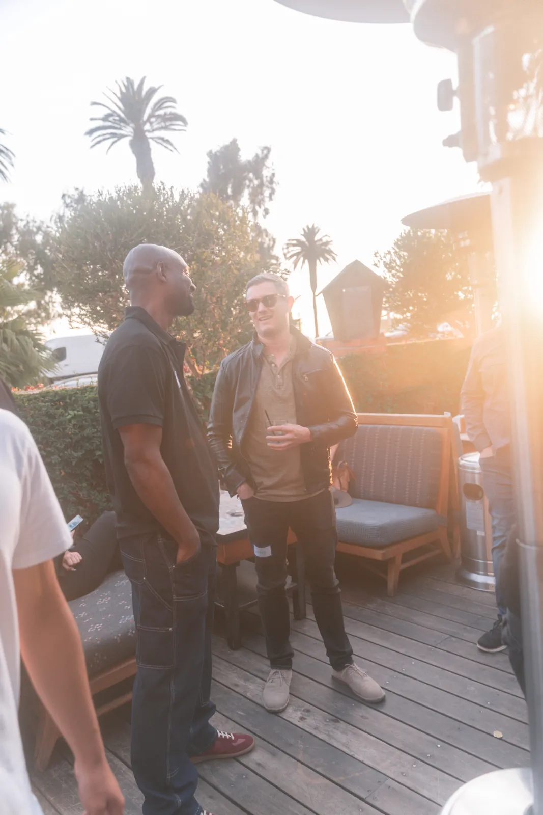 Two guests in conversation by the lounge seating as the sun flares low through the palms.