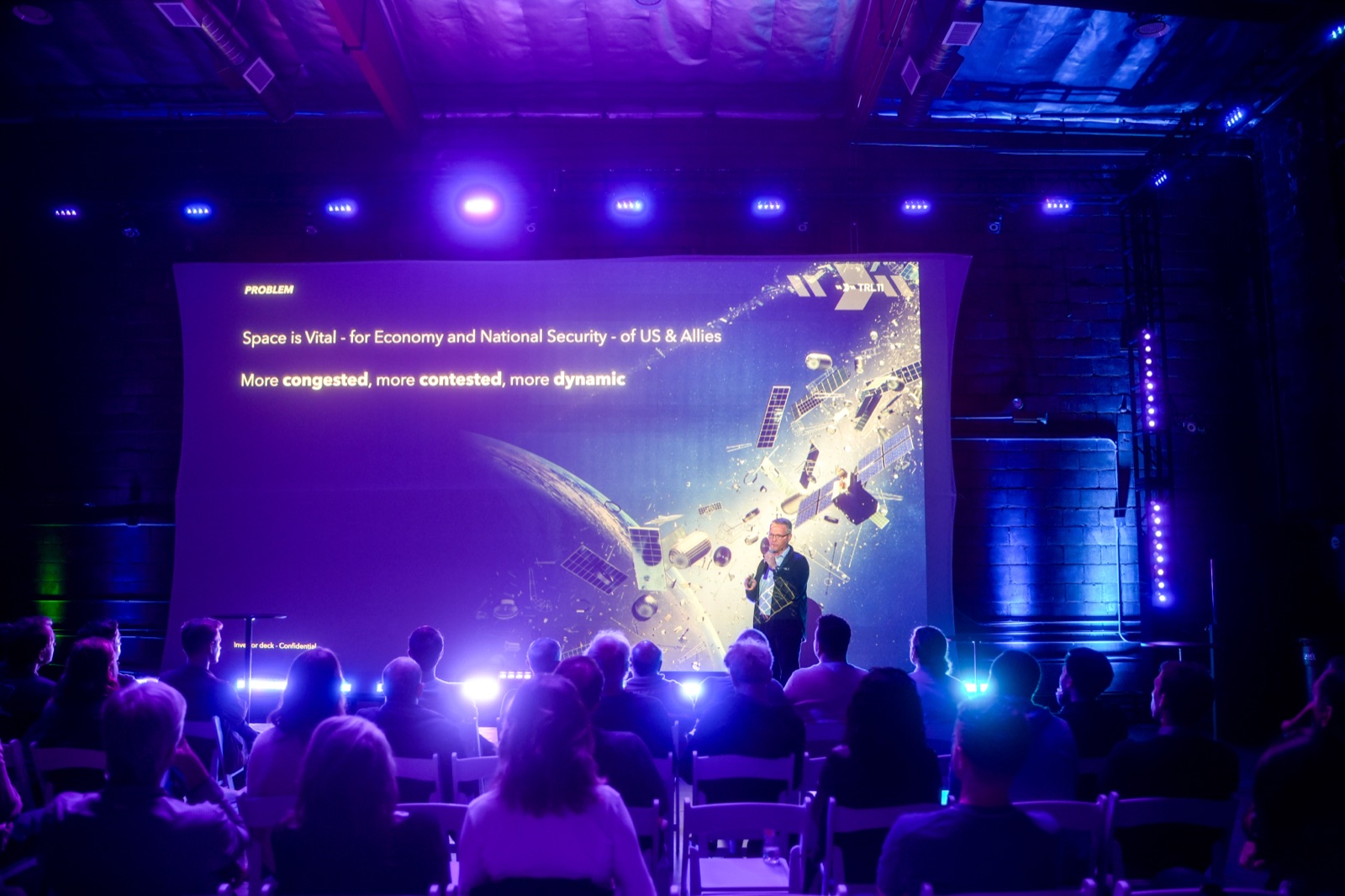 Founder pitching TRL11 on stage at Startup Arena, satellite imagery projected behind as a packed audience watches