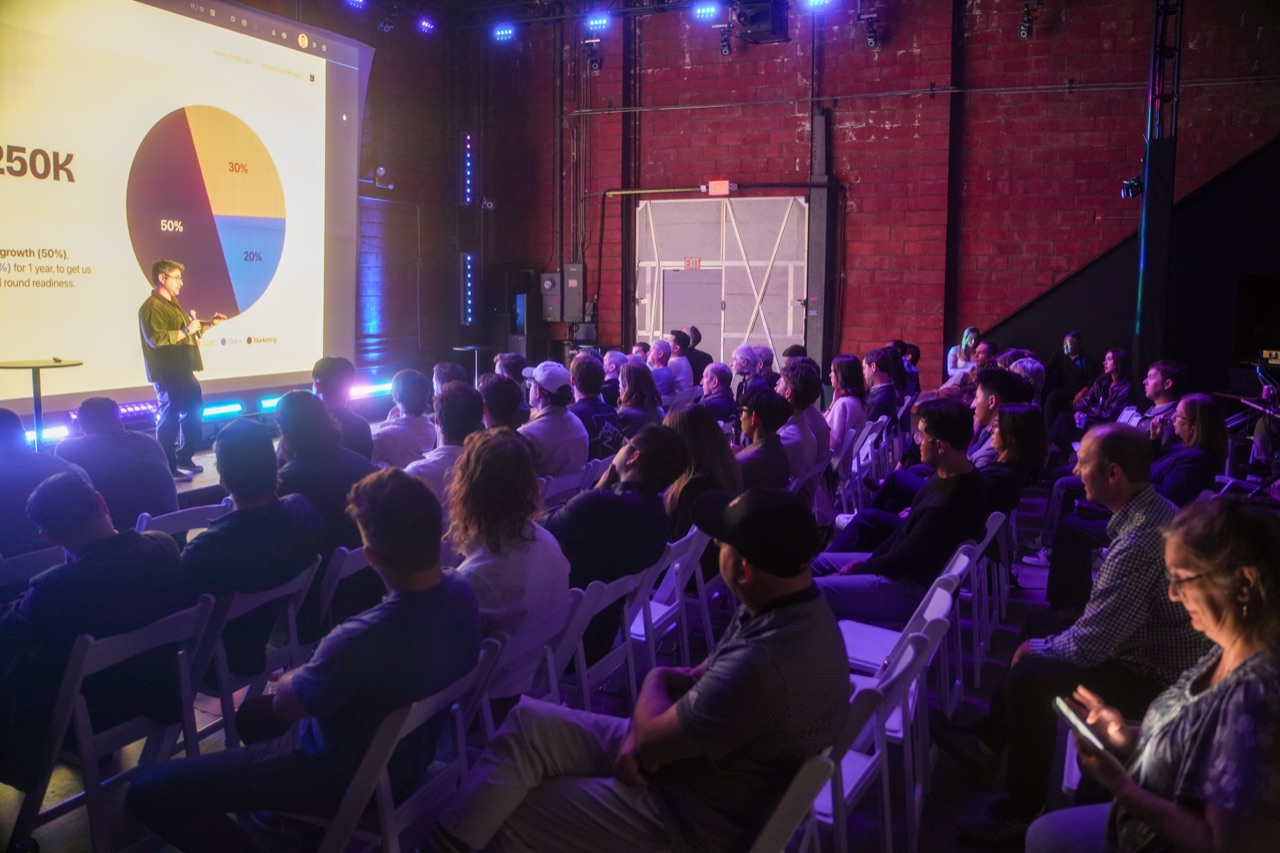 Founder pitching a pie-chart projection on stage as the audience watches from tight rows of chairs