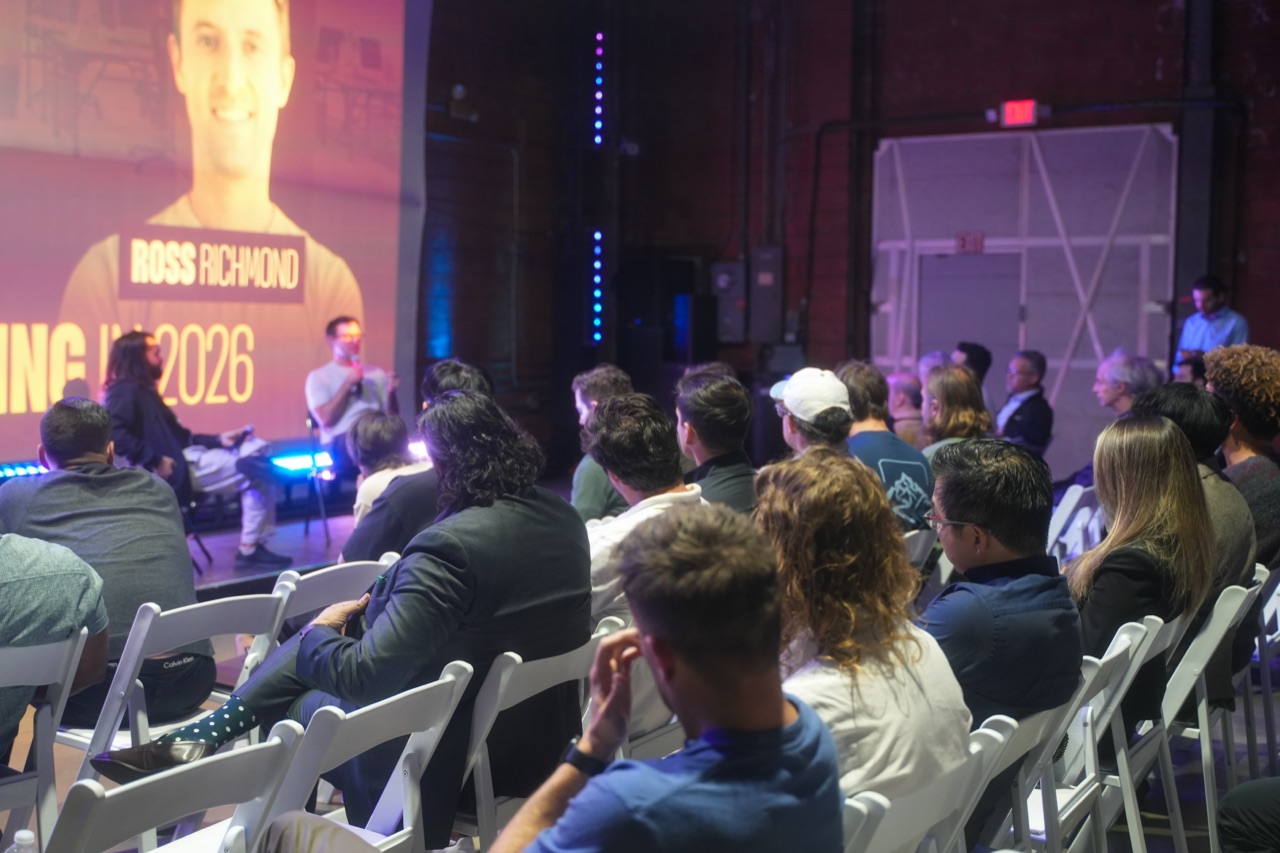 Wide shot of Ross Richmond on stage during a fireside chat, headshot projected behind, audience watching