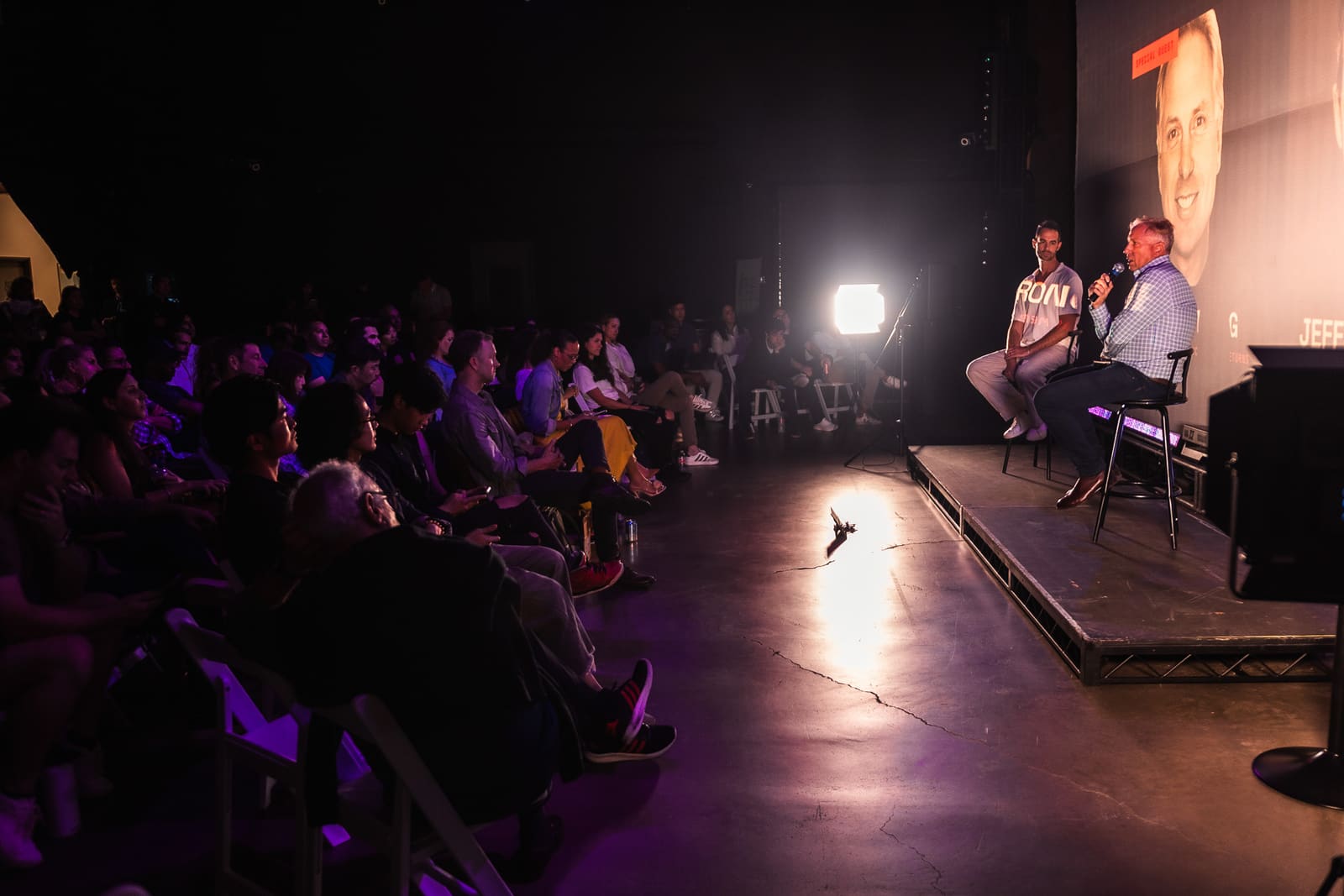 Wide view of the darkened venue as a full crowd watches Jim Armstrong and Jeffrey Chernick on the lit stage beneath the projected fireside-chat screen