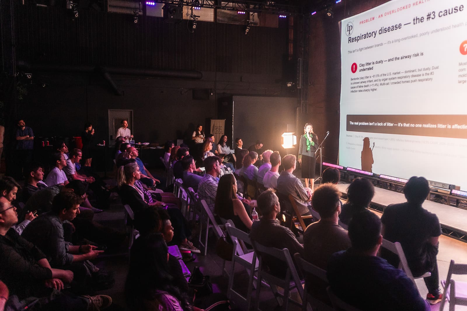 Founder pitching at a mic stand beside a giant projected slide as a packed seated audience looks on under purple rig lights
