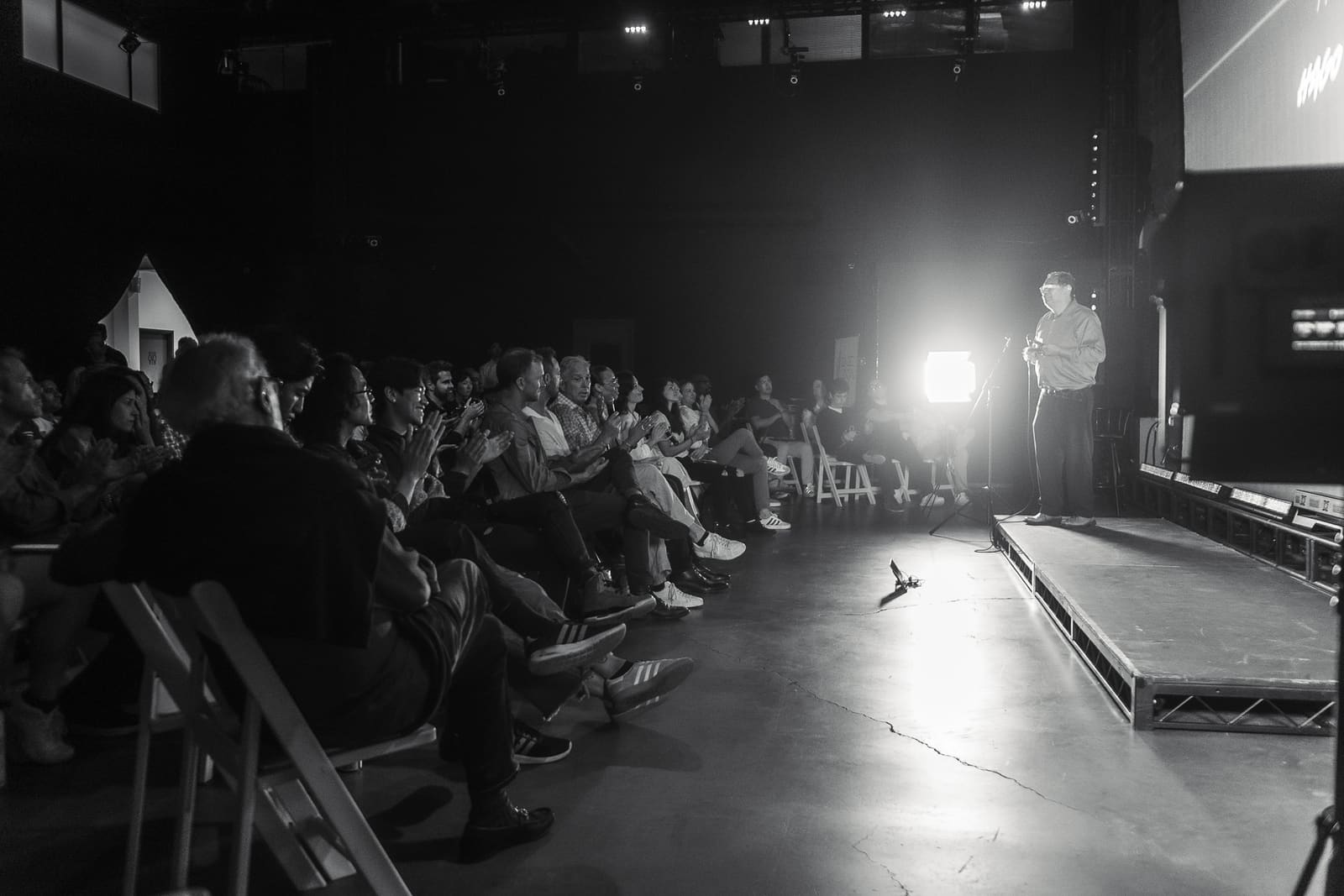 Wilson Hago standing on stage under a single bright light as the seated audience applauds in the darkened venue