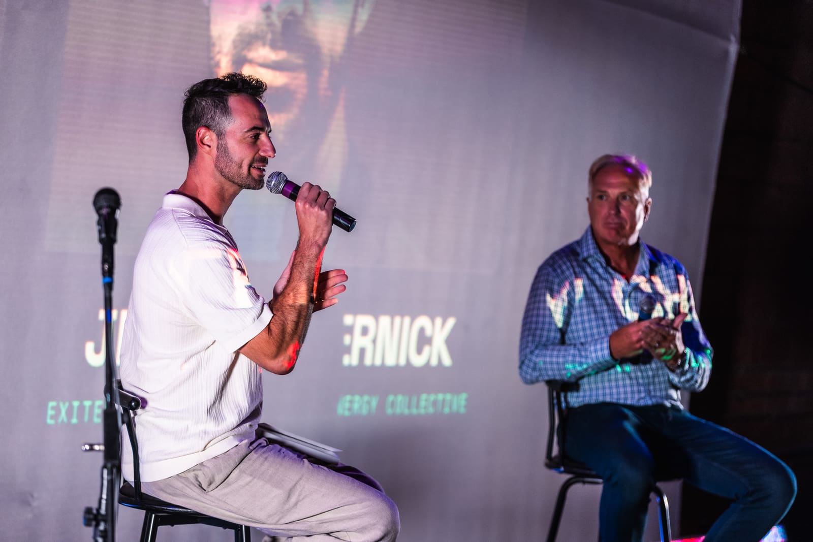 Jeffrey Chernick speaking into a mic on a stool while Jim Armstrong listens beside him, name graphics projected behind