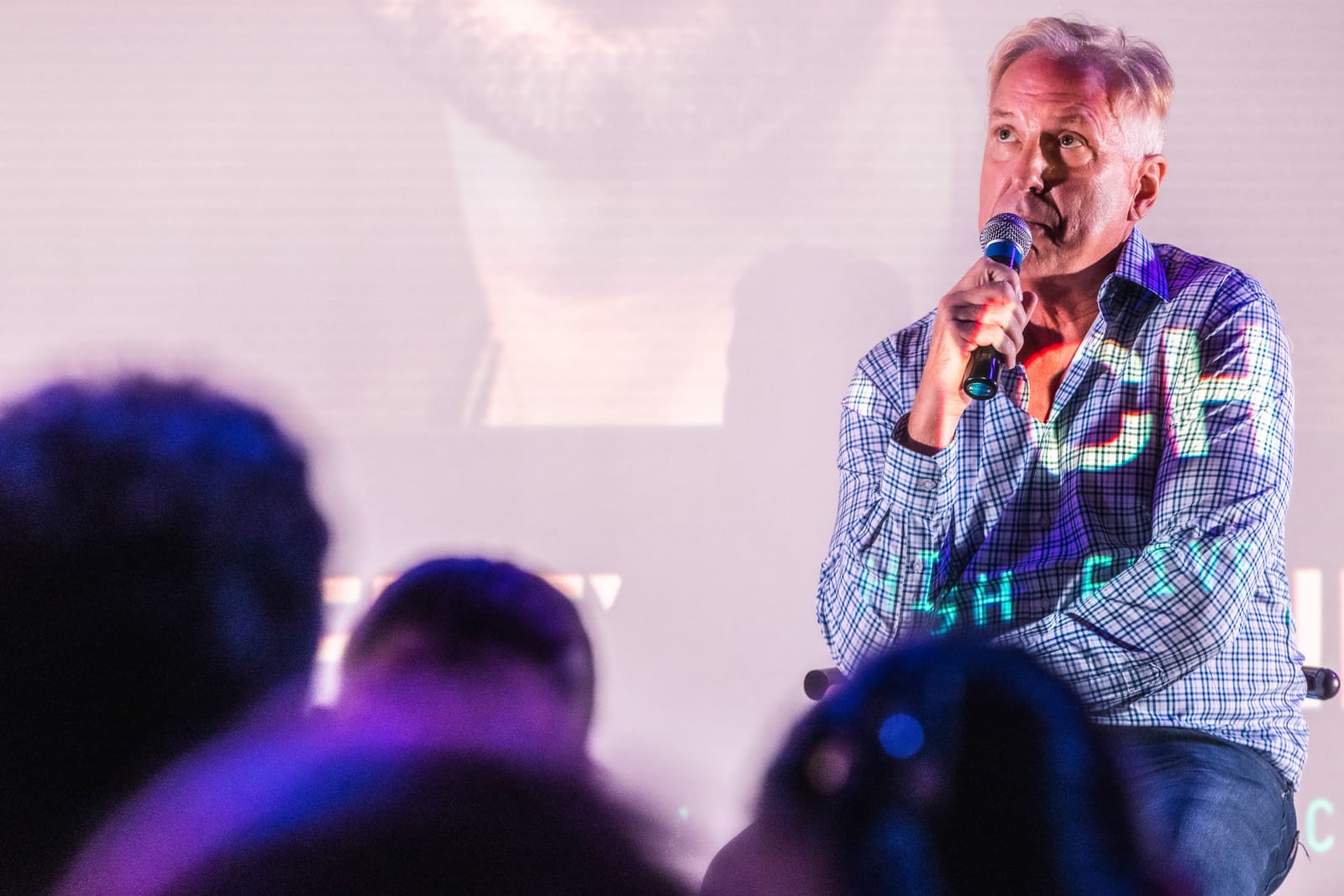 Close shot of Jim Armstrong answering into a mic, projection light dappling his shirt, audience heads blurred in the foreground