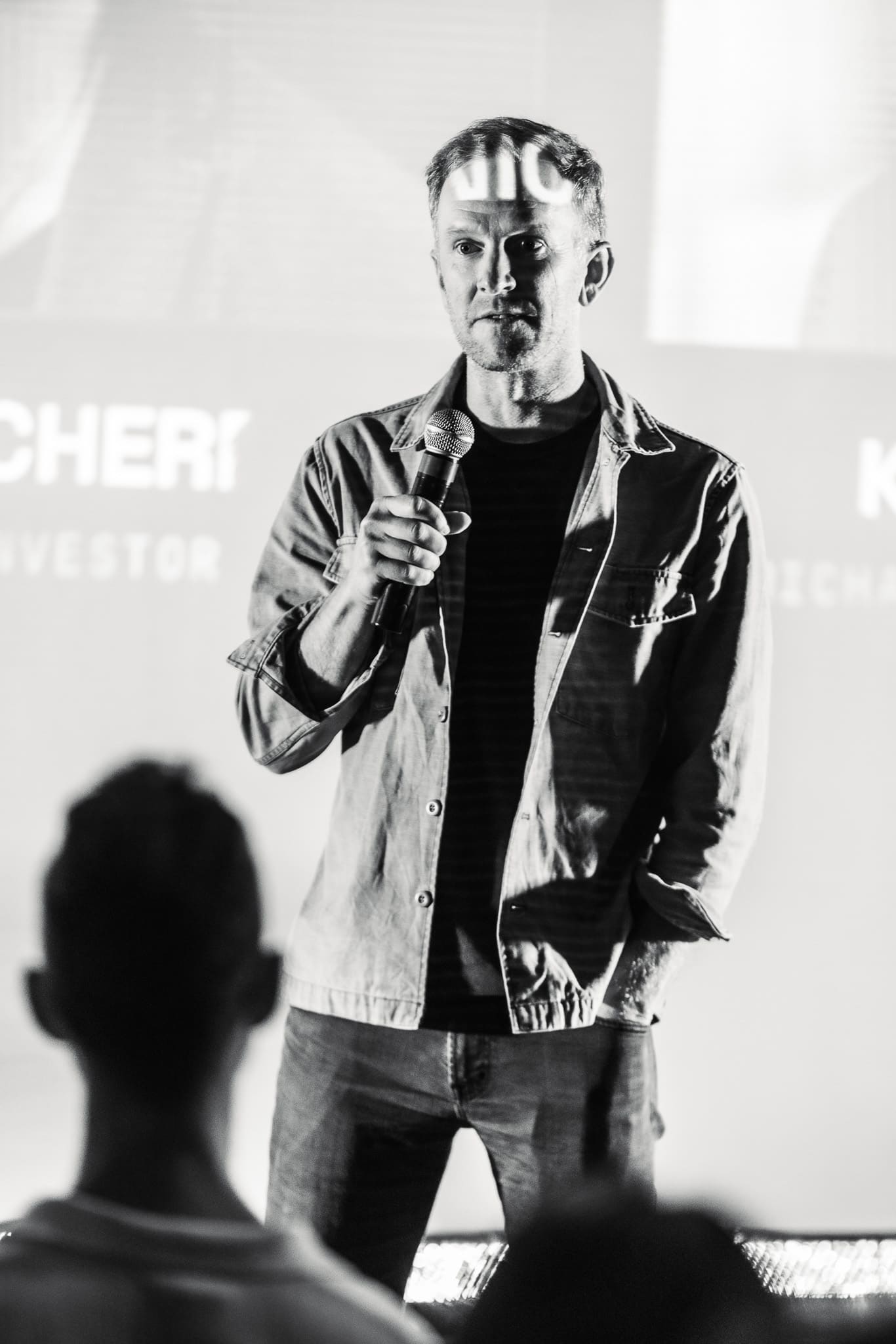 Speaker holding a mic on stage in front of a projected screen, audience silhouetted in the foreground