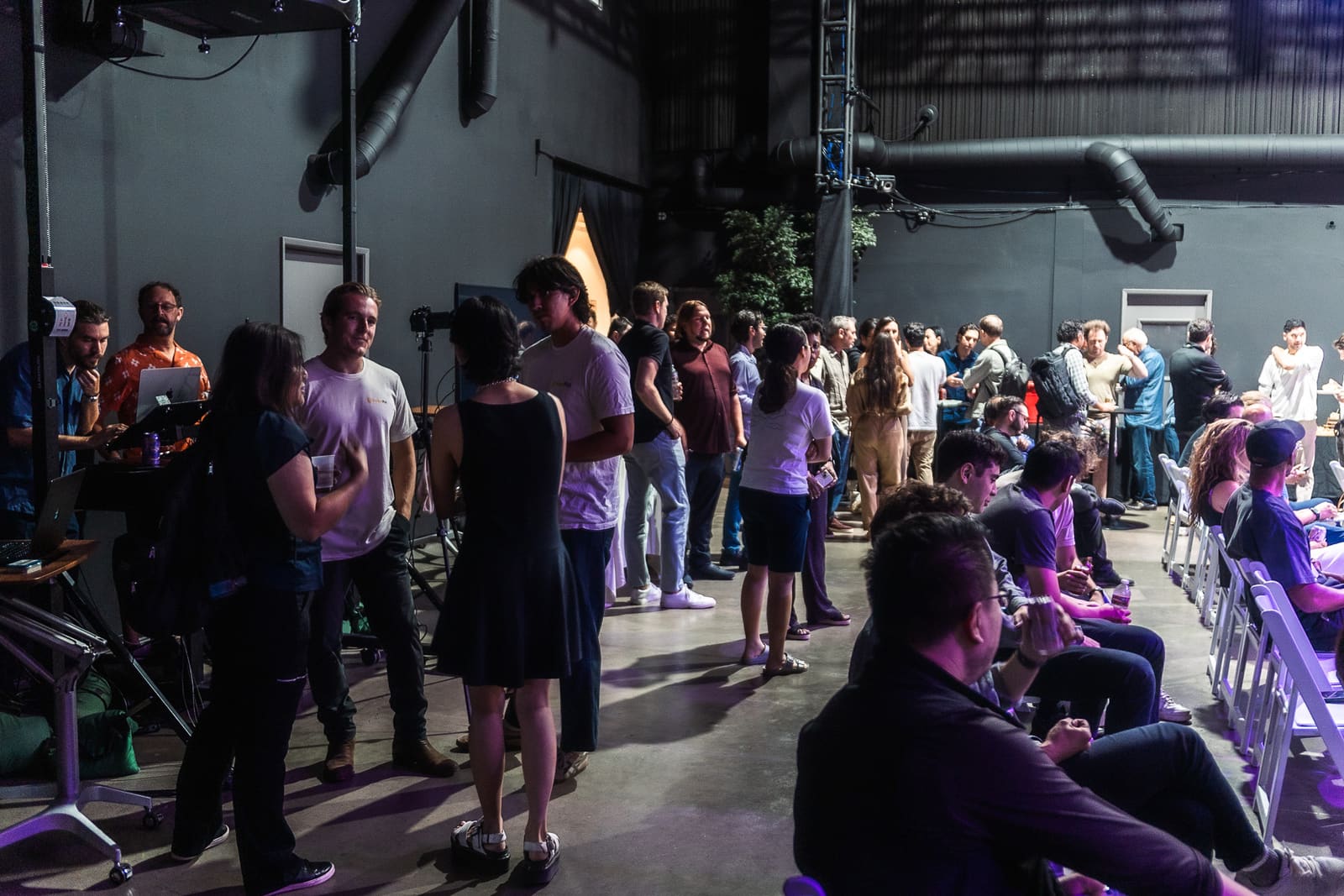The whole room mingling after the pitches, standing groups along one wall and people seated in rows of chairs