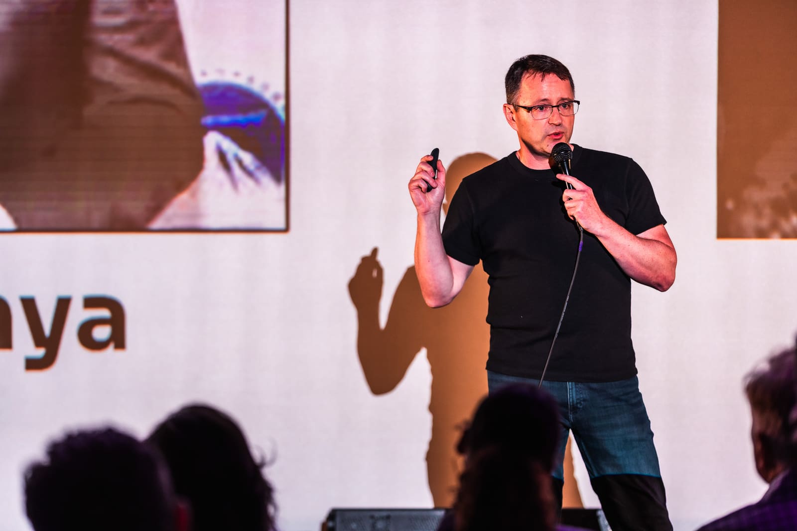 Ryan Damm of Aina pitching in a black tee, mic in one hand and clicker raised in the other, his shadow cast on the projected slide