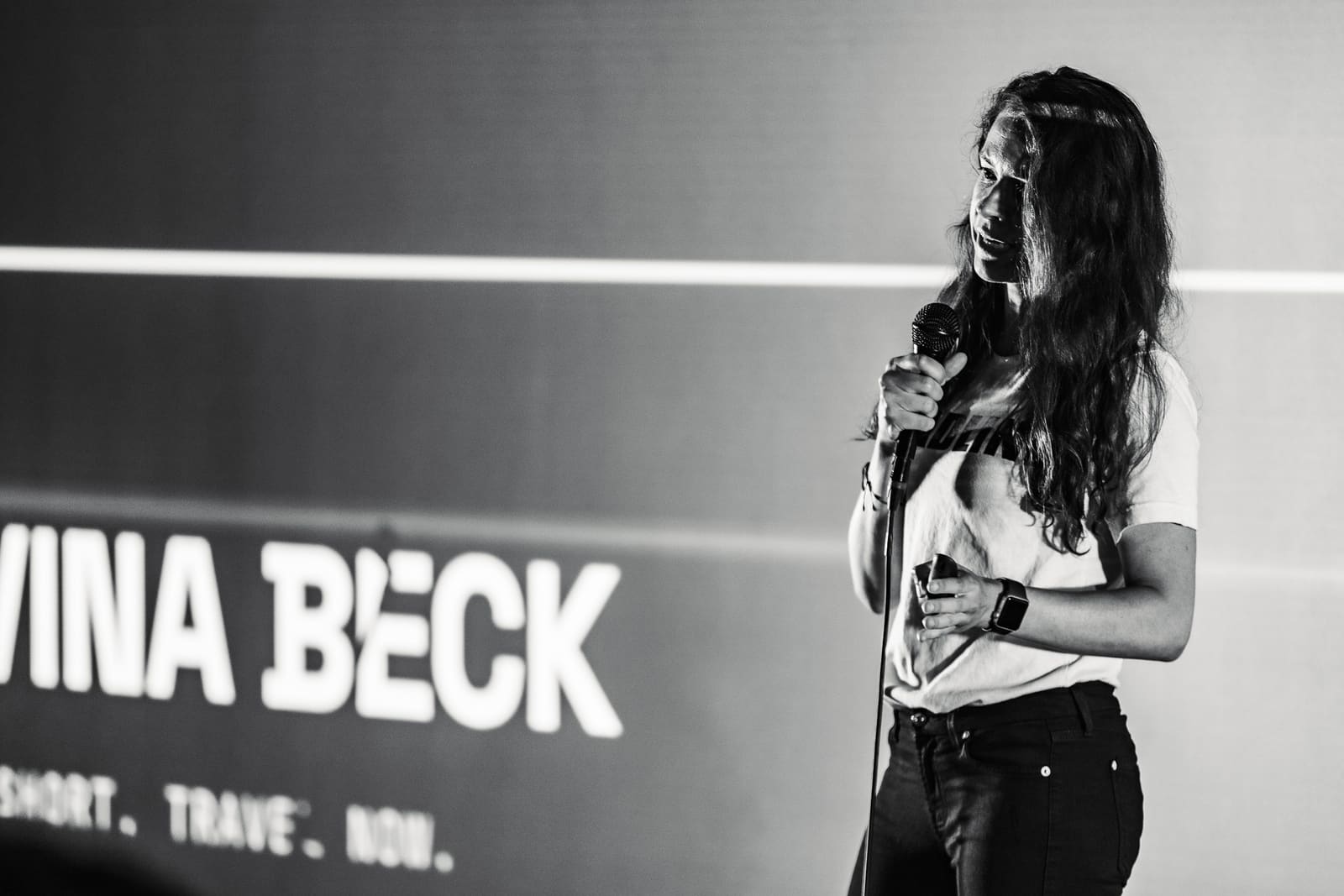Elvina Beck of Deadline speaking into a mic on stage in front of a projected screen showing her name