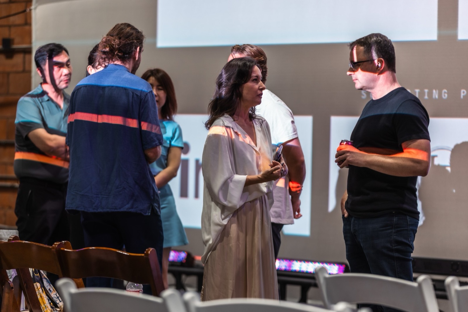 Attendees talking in small groups in front of a projection screen, stage light falling across them